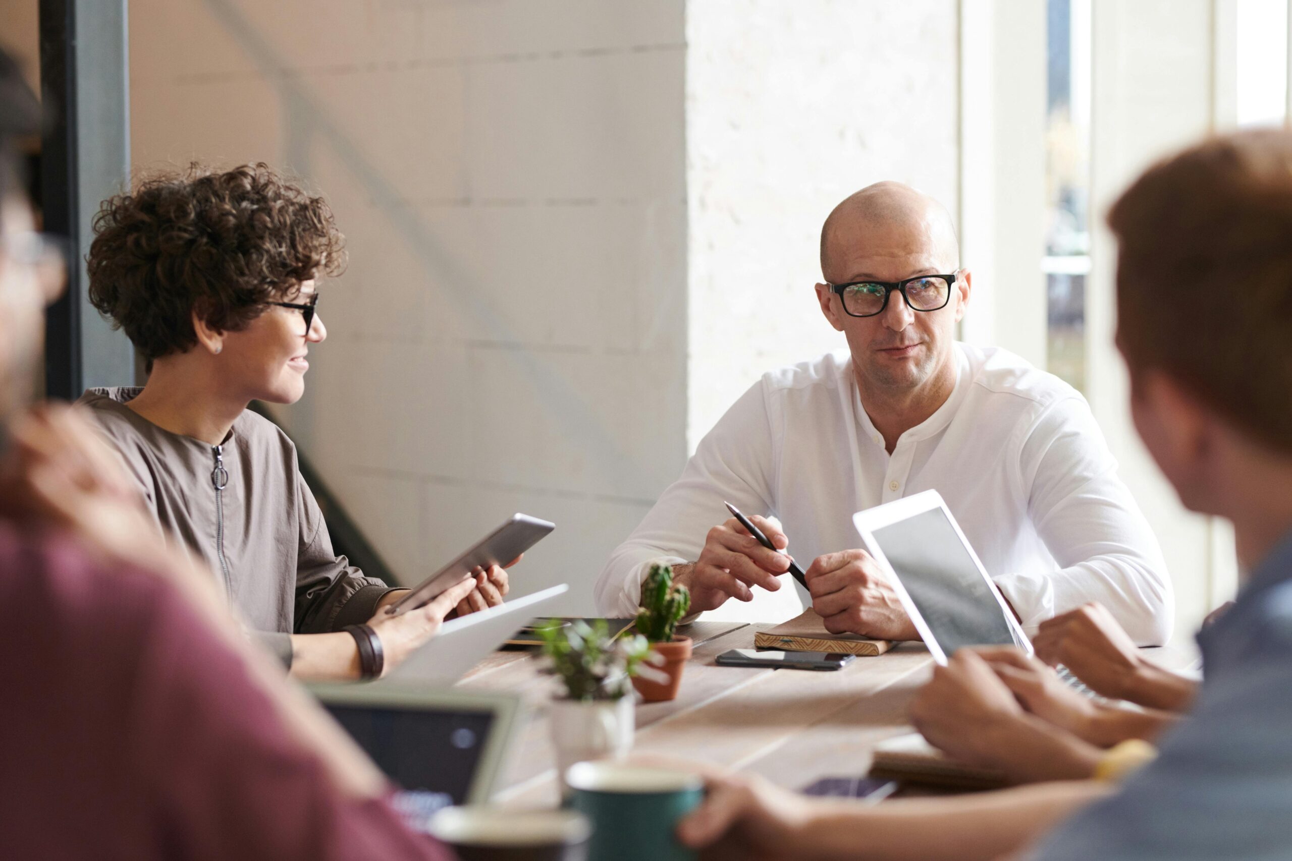 A group of colleagues seated around a wooden table in a casual meeting environment. A man in glasses and a white shirt is leading the discussion, while others listen attentively and use devices such as tablets and laptops. Small potted plants are on the table, adding a touch of greenery.