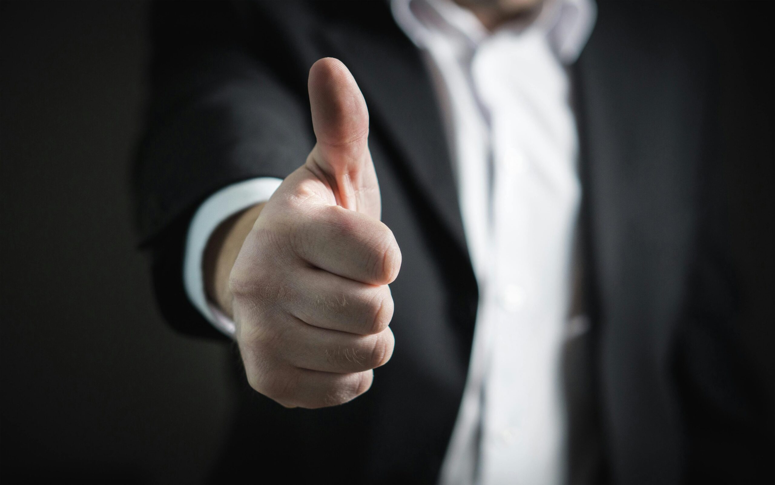 A close-up of a person in a suit giving a thumbs-up gesture, symbolizing approval or success, against a dark background.