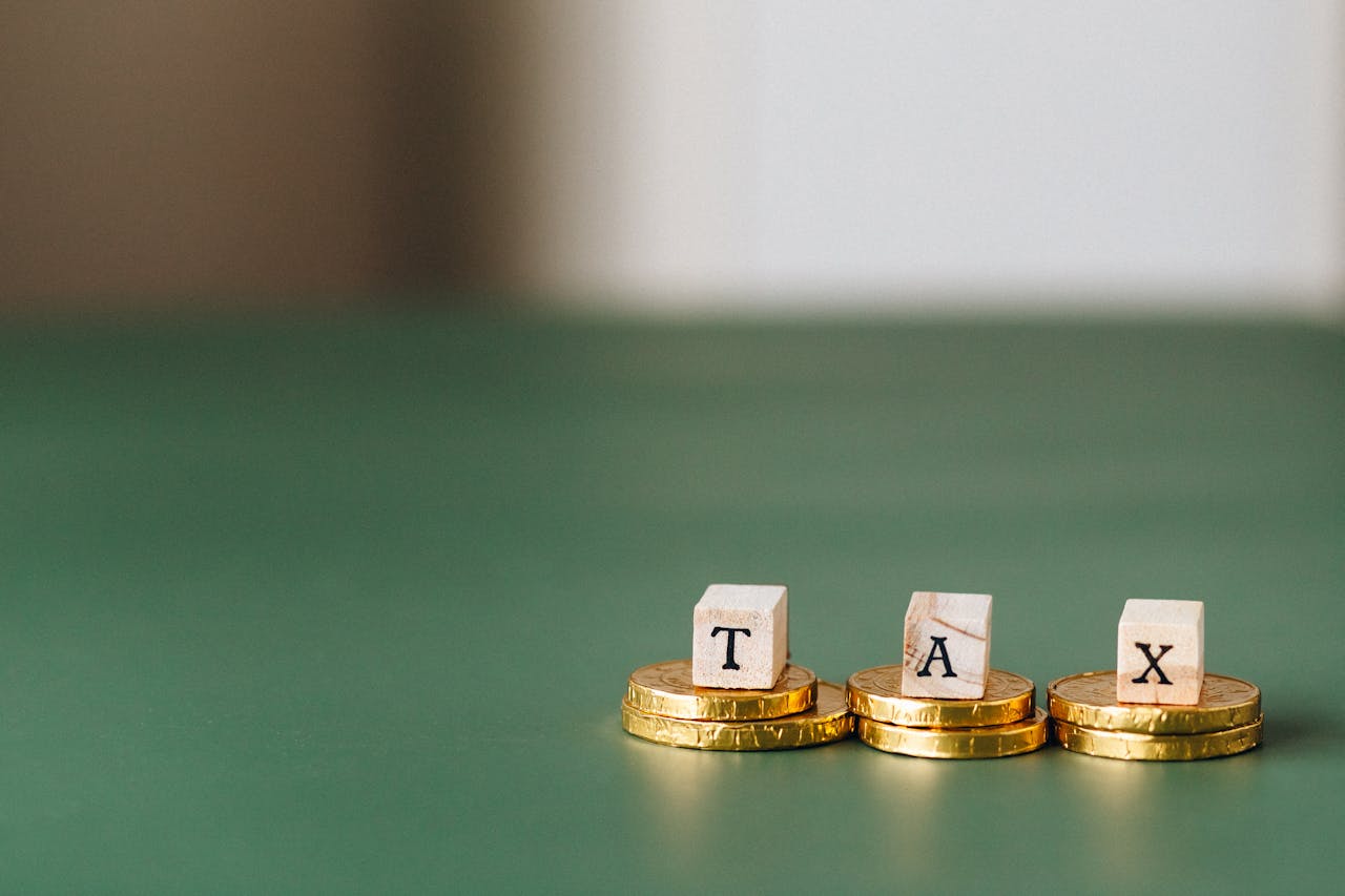 Wooden letter blocks spelling "TAX" placed on top of stacked gold coins, symbolizing financial taxation or economic management. The background is a smooth, green surface with blurred depth.